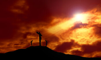 A depiction of the crucifixion of Jesus Christ on a cross with 2 other robbers against a dramatic sunset with rays of light breaking through the clouds onto the cross and lens flare for effect. Concept of the death of Jesus on Good Friday and His resurrection on Easter Sunday. Horizontal orientation with copy space.